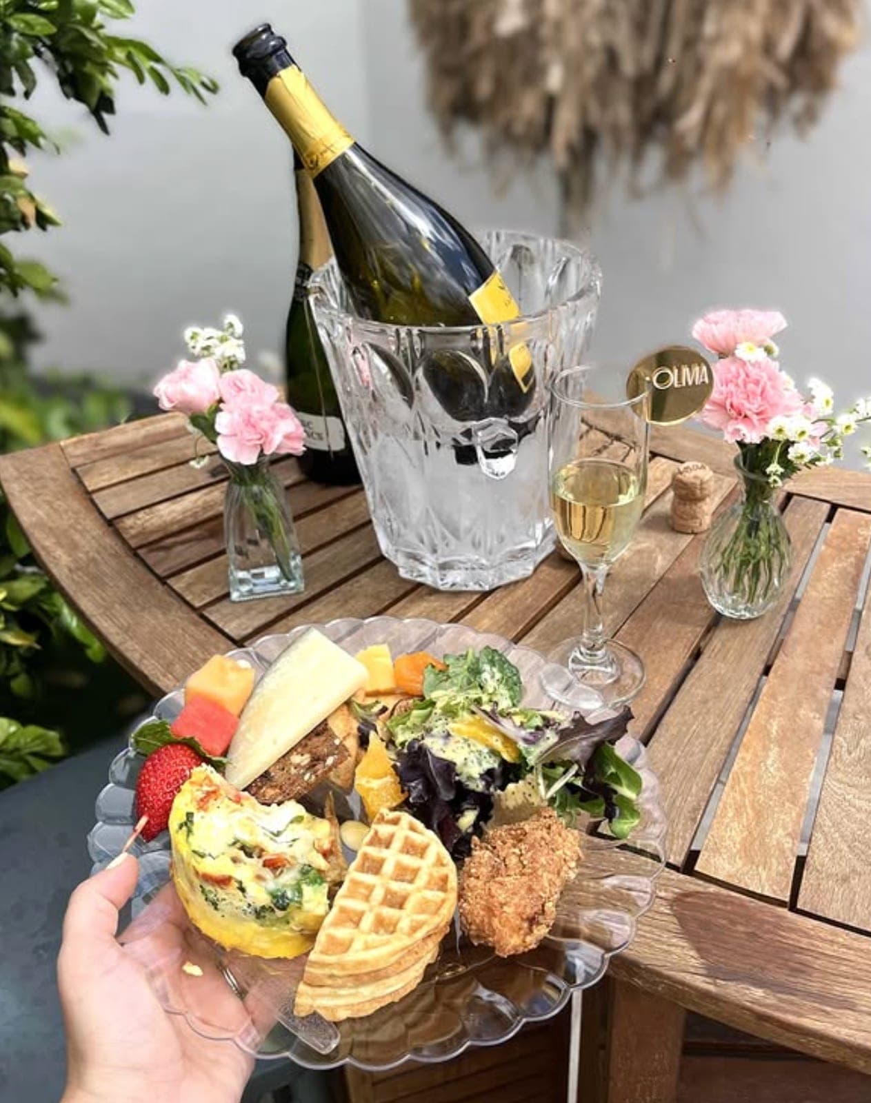 A plated celebration brunch — frittata, waffle, fried chicken, salad, and fruit — with champagne and florals