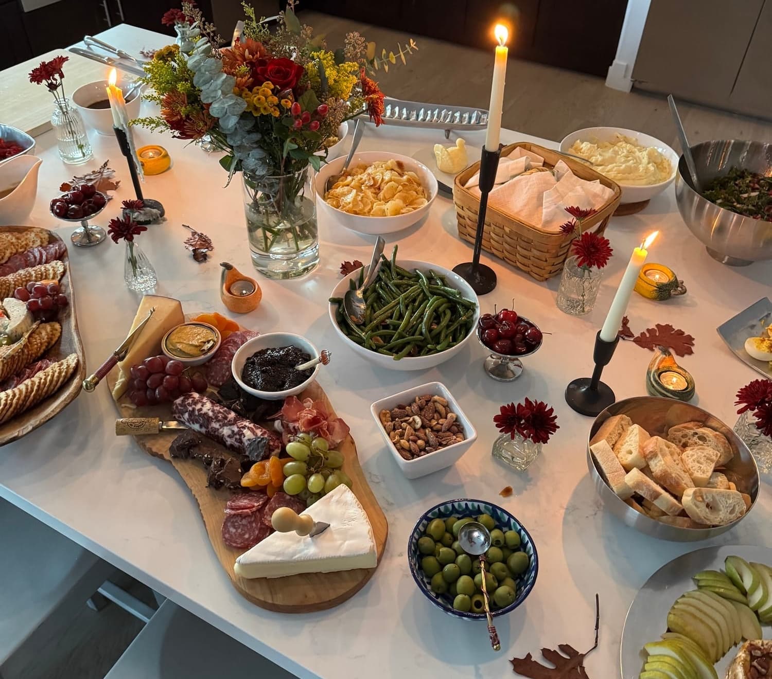 Overhead view of a holiday spread on a marble island — charcuterie, mashed potatoes, green beans, olives, breads, and fall florals
