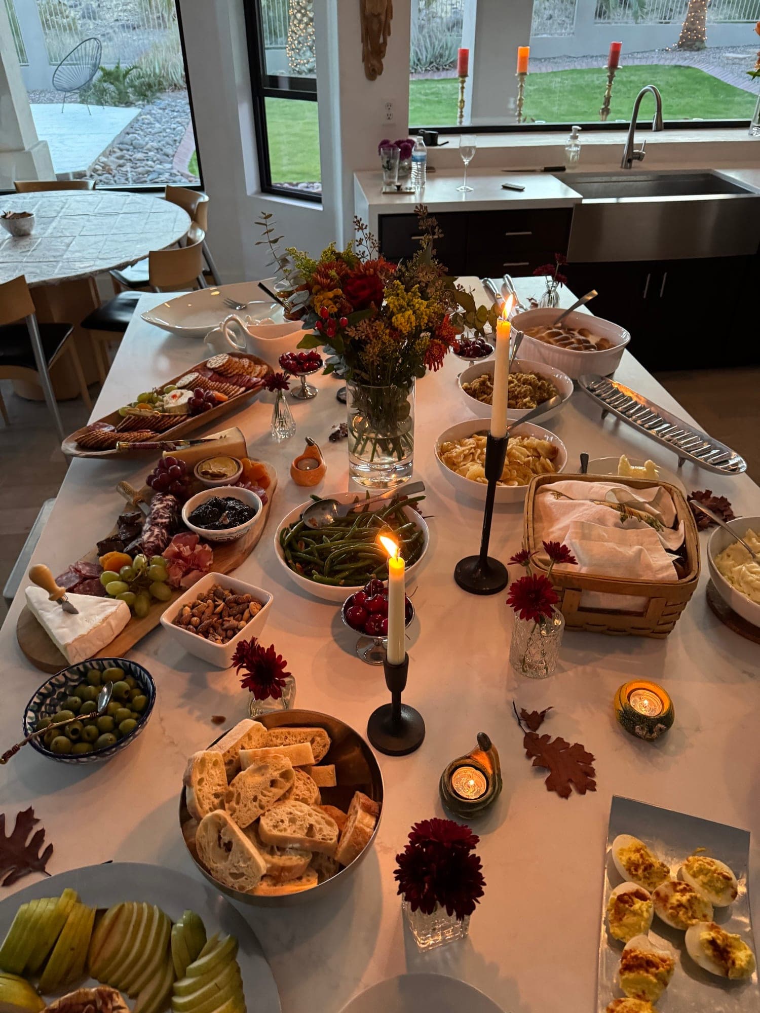 A candlelit holiday spread on a marble island — charcuterie, breads, sides, and fall florals