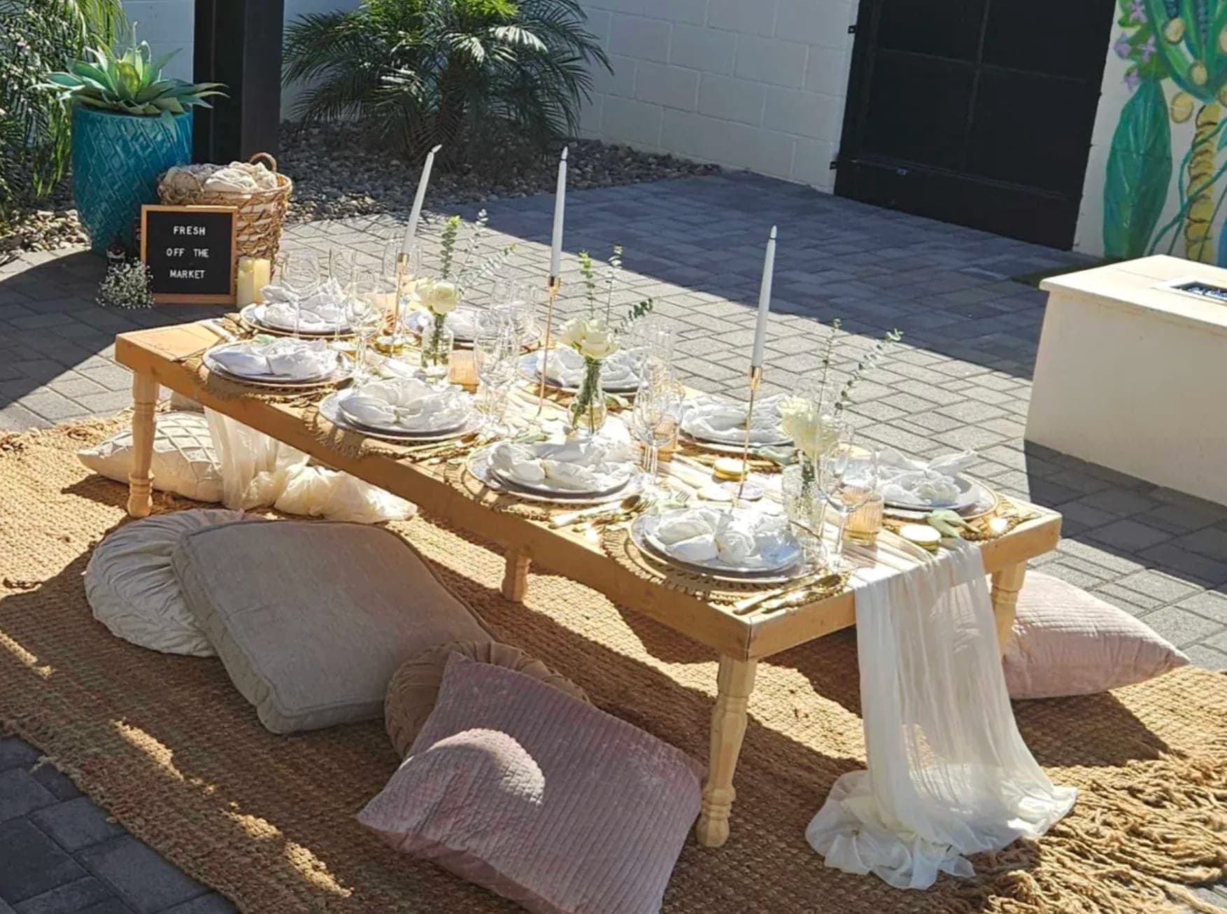 A styled low picnic table with rental place settings, glassware, candlesticks, floor cushions, and jute rugs